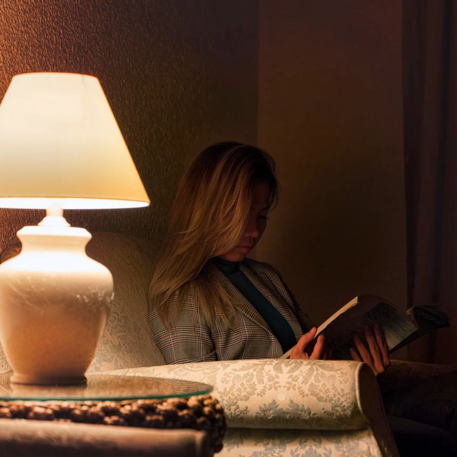 Eine junge Frau liest ein Buch auf dem Sofa. Im Vordergrund ist eine Lampe zu sehen. Die Frau sitzt im Halbschatten